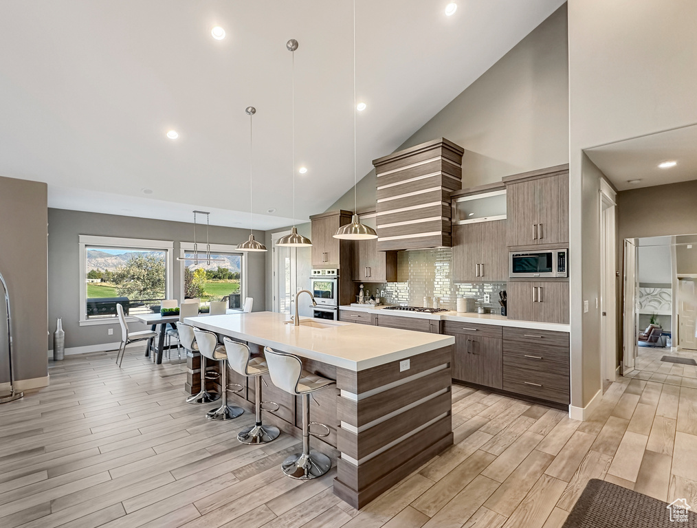 Kitchen featuring light wood-type flooring, high vaulted ceiling, a center island with sink, appliances with stainless steel finishes, and decorative light fixtures