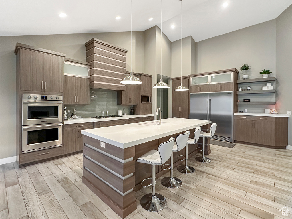 Kitchen featuring built in appliances, pendant lighting, light wood-type flooring, a center island with sink, and sink