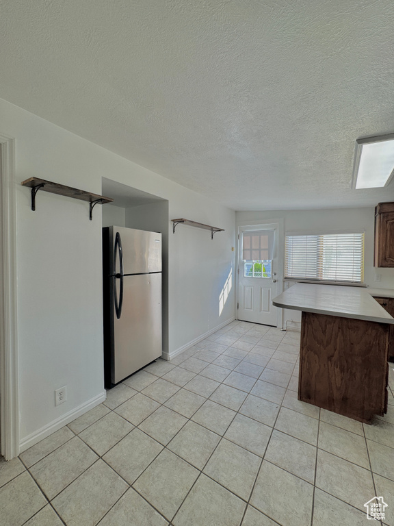 Kitchen with a textured ceiling, kitchen peninsula, stainless steel refrigerator, dark brown cabinetry, and light tile patterned floors