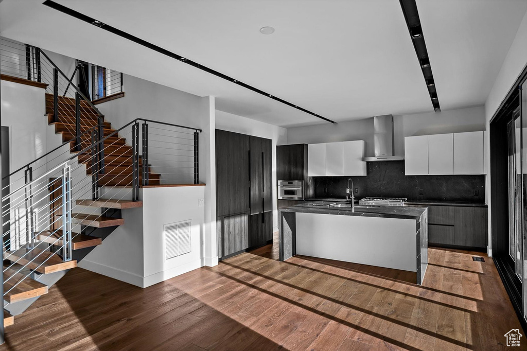 Kitchen with tasteful backsplash, wood-type flooring, wall chimney range hood, white cabinets, and a kitchen island with sink