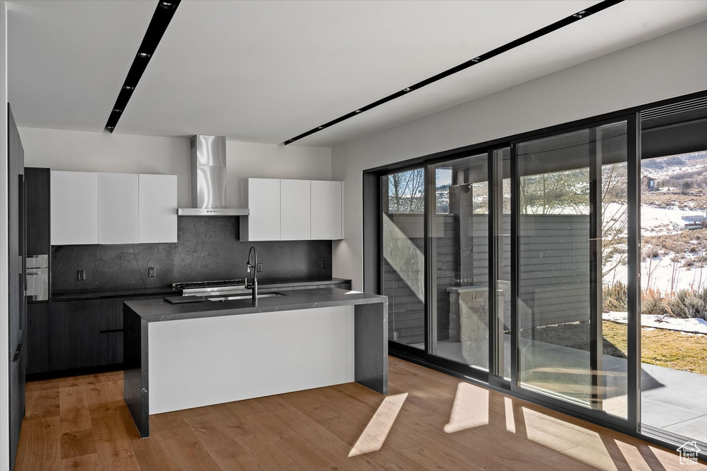 Kitchen with wall chimney range hood, tasteful backsplash, hardwood / wood-style floors, an island with sink, and white cabinets