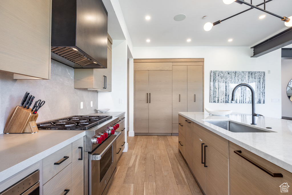 Kitchen with light brown cabinets, wall chimney range hood, sink, light hardwood / wood-style flooring, and stainless steel range