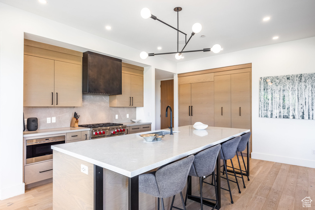 Kitchen featuring light brown cabinets, wall chimney exhaust hood, backsplash, decorative light fixtures, and a kitchen island with sink