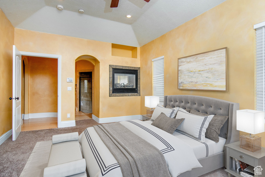 Carpeted bedroom featuring ceiling fan, a textured ceiling, a multi sided fireplace, and vaulted ceiling