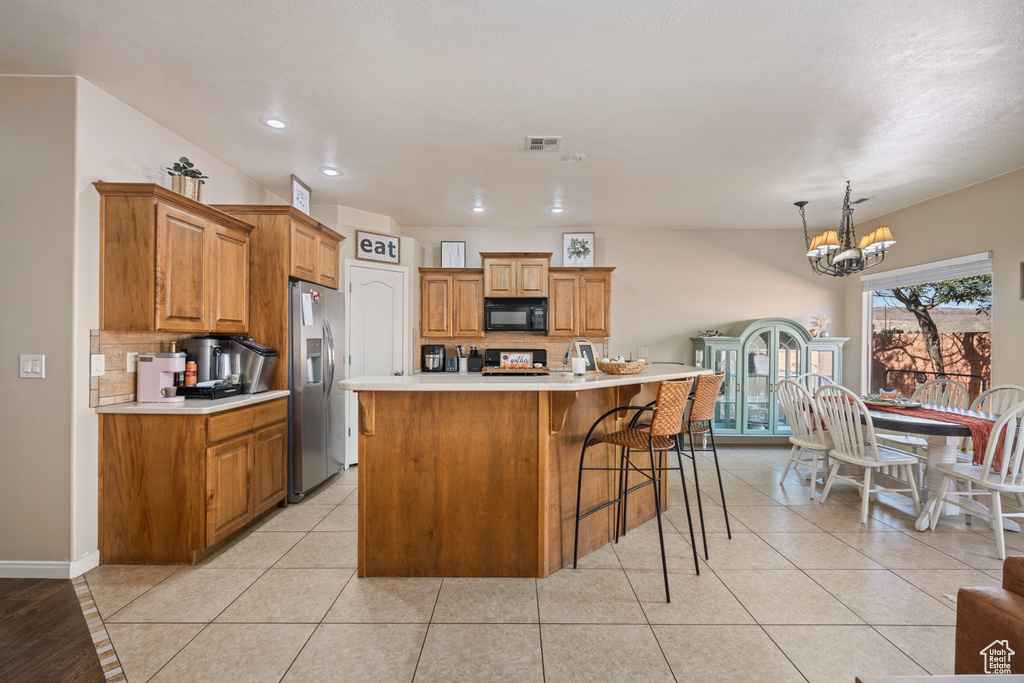 Kitchen with pendant lighting, stainless steel fridge, a kitchen island with sink, backsplash, and a notable chandelier