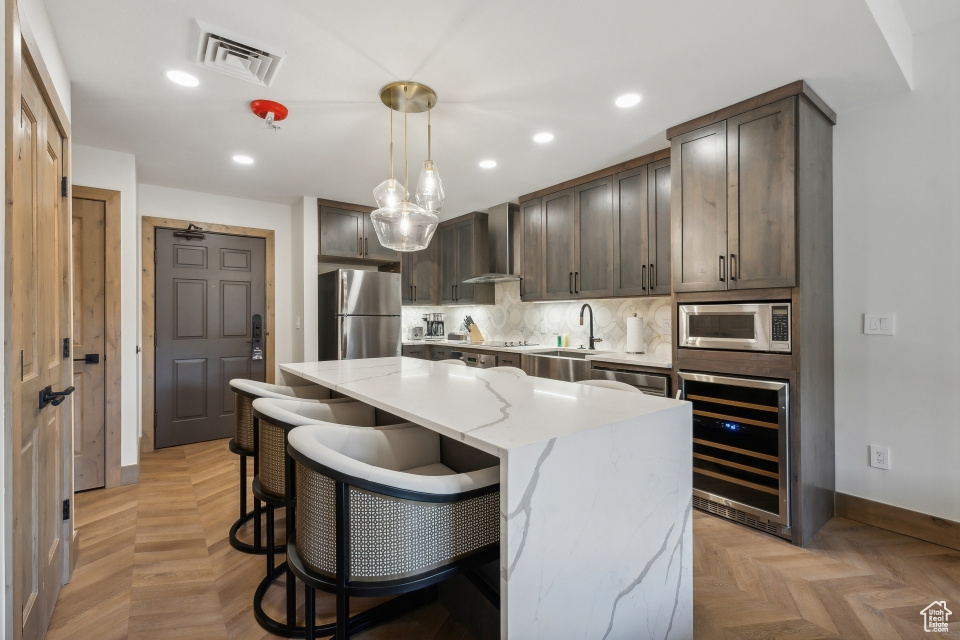 Kitchen featuring wine cooler, light stone counters, a kitchen island, stainless steel appliances, and wall chimney range hood