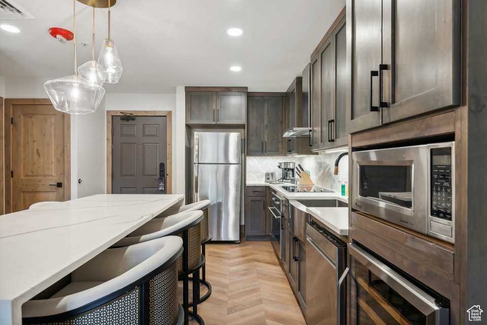 Kitchen with decorative backsplash, light parquet flooring, appliances with stainless steel finishes, decorative light fixtures, and light stone countertops