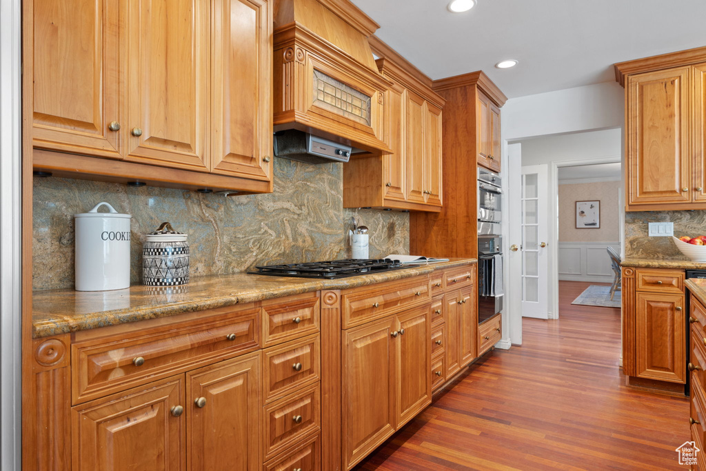 Kitchen with tasteful backsplash, dark wood-type flooring, stainless steel appliances, custom range hood, and light stone countertops