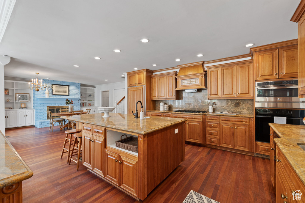 Kitchen with premium range hood, dark wood-type flooring, pendant lighting, stainless steel double oven, and a center island with sink