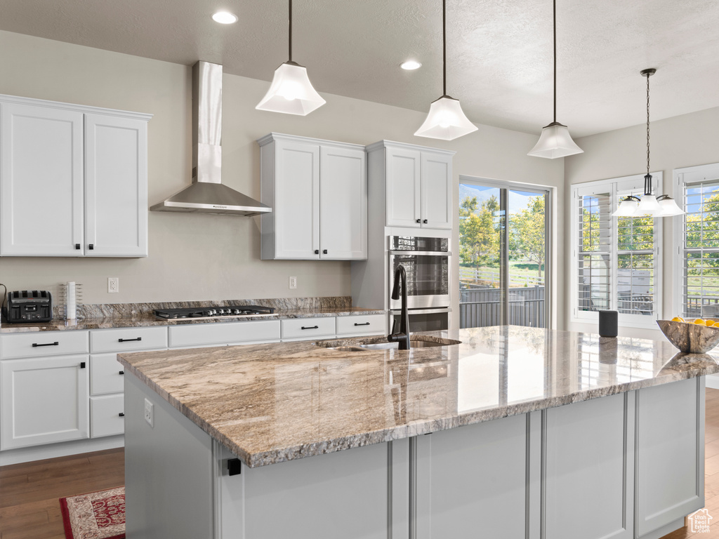 Kitchen featuring a center island with sink, wall chimney exhaust hood, white cabinetry, and decorative light fixtures