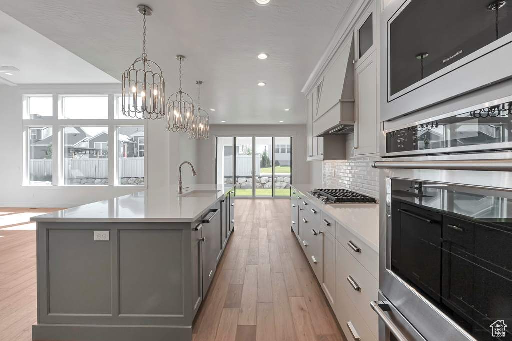 Kitchen featuring light wood-type flooring, a kitchen island with sink, stainless steel gas stovetop, white cabinetry, and an inviting chandelier