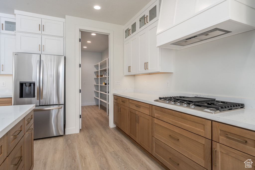 Kitchen featuring white cabinets, custom exhaust hood, light hardwood / wood-style flooring, appliances with stainless steel finishes, and light stone countertops