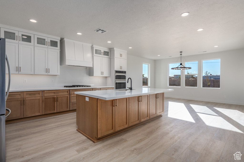 Kitchen featuring appliances with stainless steel finishes, hanging light fixtures, an island with sink, white cabinets, and light hardwood / wood-style flooring