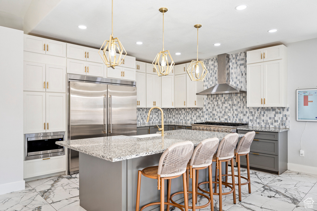 Kitchen with light stone counters, white cabinets, hanging light fixtures, a kitchen island with sink, and wall chimney exhaust hood