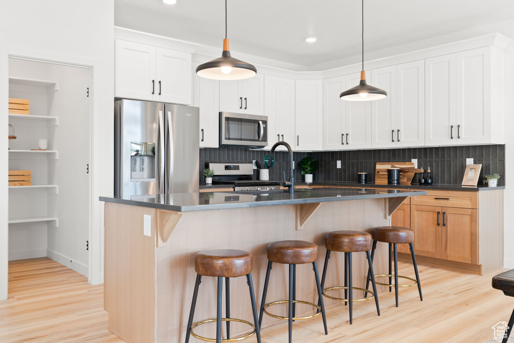 Kitchen with white cabinets, a kitchen island with sink, backsplash, stainless steel appliances, and light wood-type flooring