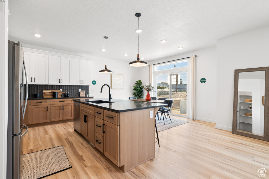 Kitchen with light hardwood / wood-style floors, white cabinetry, an island with sink, stainless steel appliances, and decorative light fixtures