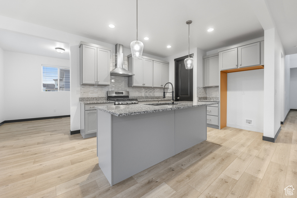 Kitchen featuring light hardwood / wood-style floors, an island with sink, wall chimney exhaust hood, decorative light fixtures, and gas stove