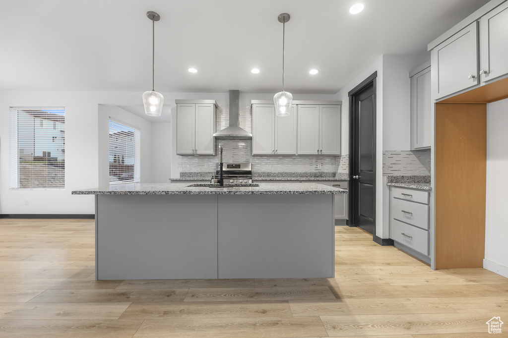 Kitchen featuring an island with sink, decorative light fixtures, wall chimney exhaust hood, light hardwood / wood-style flooring, and light stone countertops