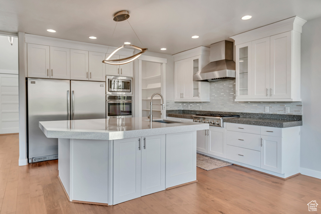 Kitchen featuring sink, backsplash, wall chimney range hood, built in appliances, and light wood-type flooring