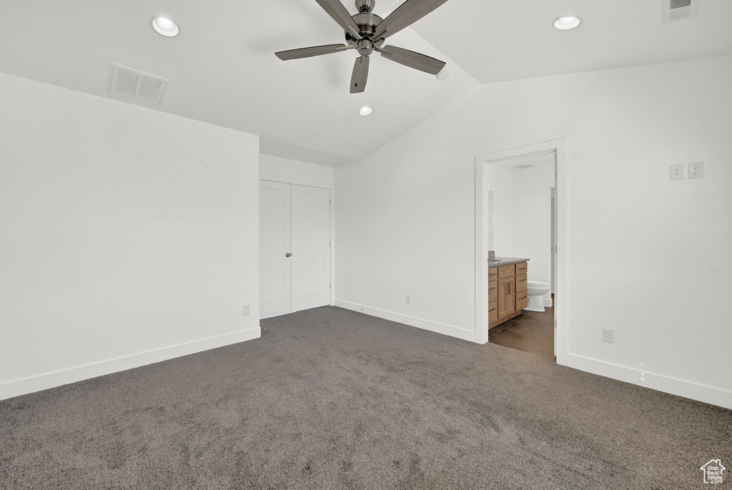 Unfurnished bedroom with lofted ceiling, dark carpet, ensuite bath, and ceiling fan