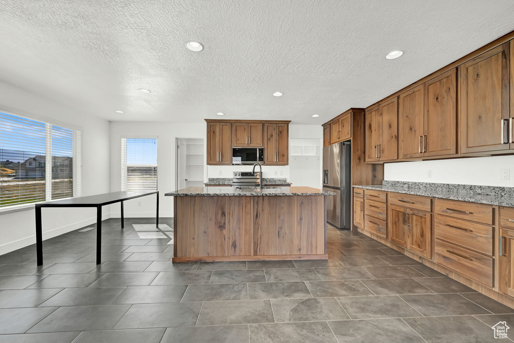 Kitchen featuring dark tile patterned flooring, a textured ceiling, a kitchen island with sink, stainless steel appliances, and dark stone countertops