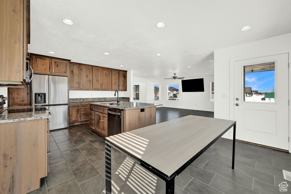 Kitchen with an island with sink, light stone counters, stainless steel appliances, ceiling fan, and sink