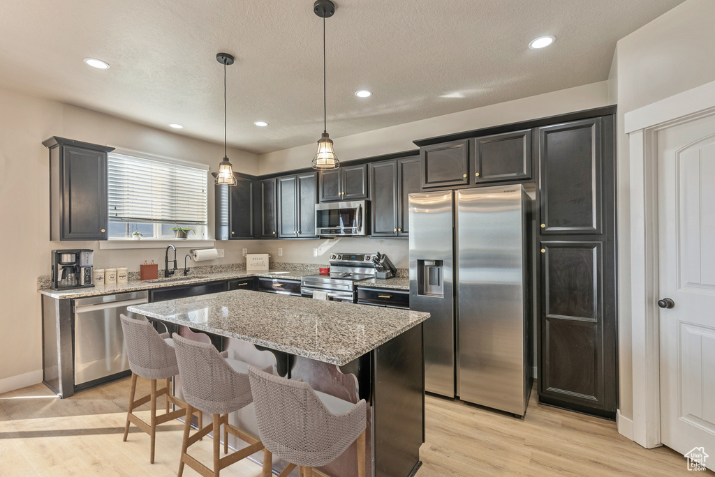 Kitchen with light hardwood / wood-style floors, a kitchen island, decorative light fixtures, light stone countertops, and appliances with stainless steel finishes