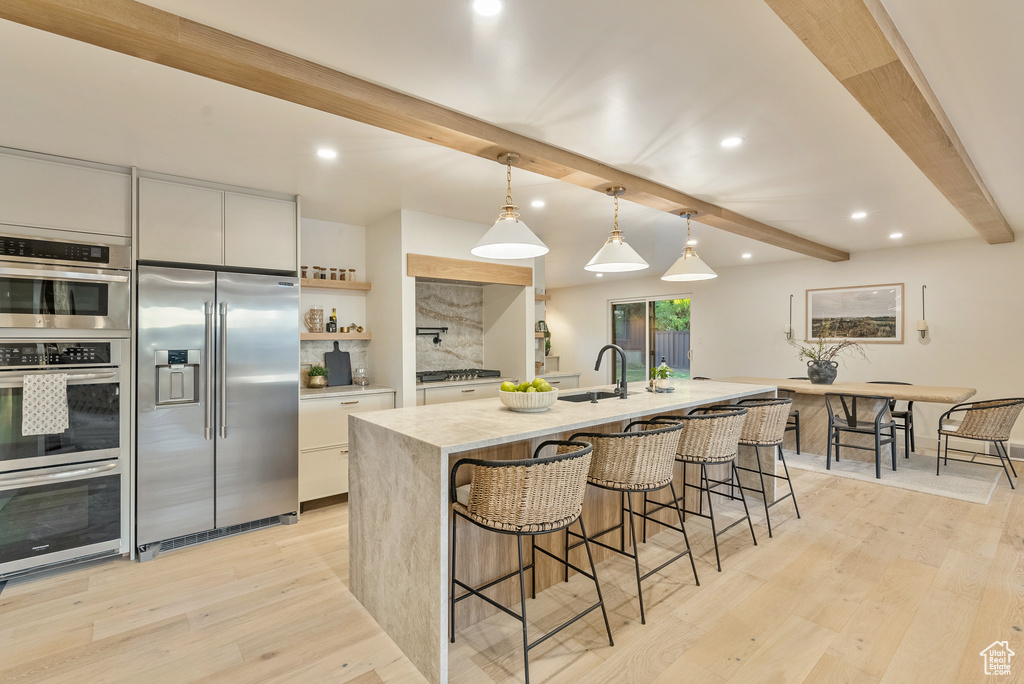 Kitchen featuring white cabinets, an island with sink, hanging light fixtures, beam ceiling, and stainless steel appliances