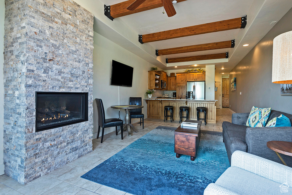 Living room with ceiling fan, a stone fireplace, light tile patterned flooring, and beam ceiling