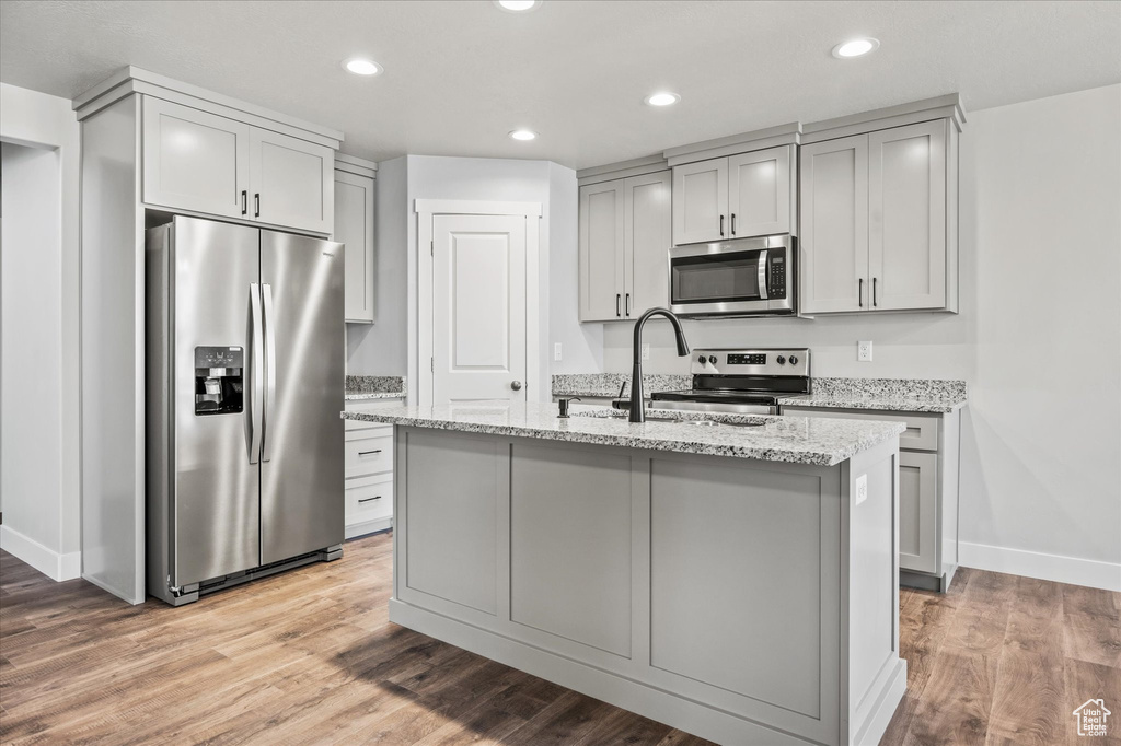 Kitchen featuring light hardwood / wood-style flooring, sink, stainless steel appliances, light stone countertops, and an island with sink