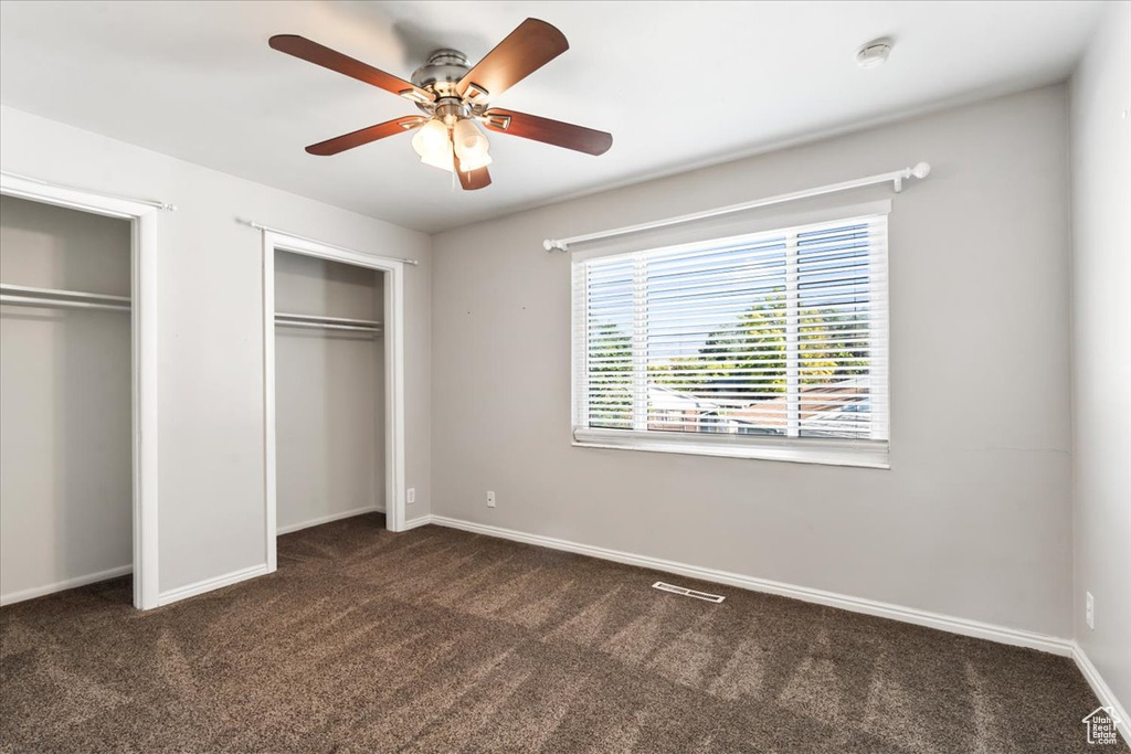 Unfurnished bedroom with ceiling fan, dark carpet, and two closets