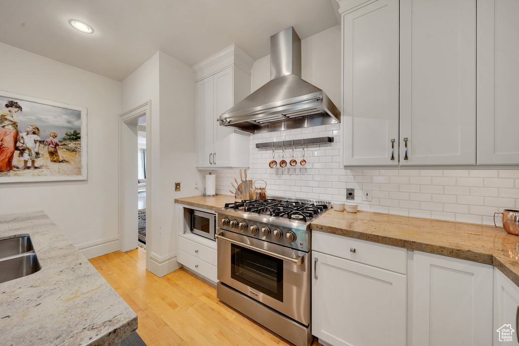 Kitchen with wall chimney exhaust hood, white cabinets, appliances with stainless steel finishes, light stone counters, and light hardwood / wood-style floors