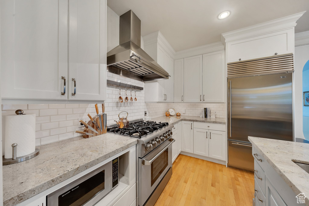 Kitchen featuring built in appliances, wall chimney range hood, light wood-type flooring, and white cabinetry