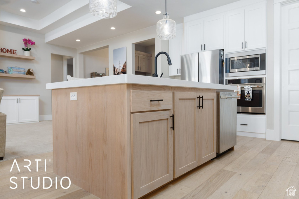 Kitchen featuring light brown cabinets, light hardwood / wood-style floors, appliances with stainless steel finishes, a center island with sink, and decorative light fixtures