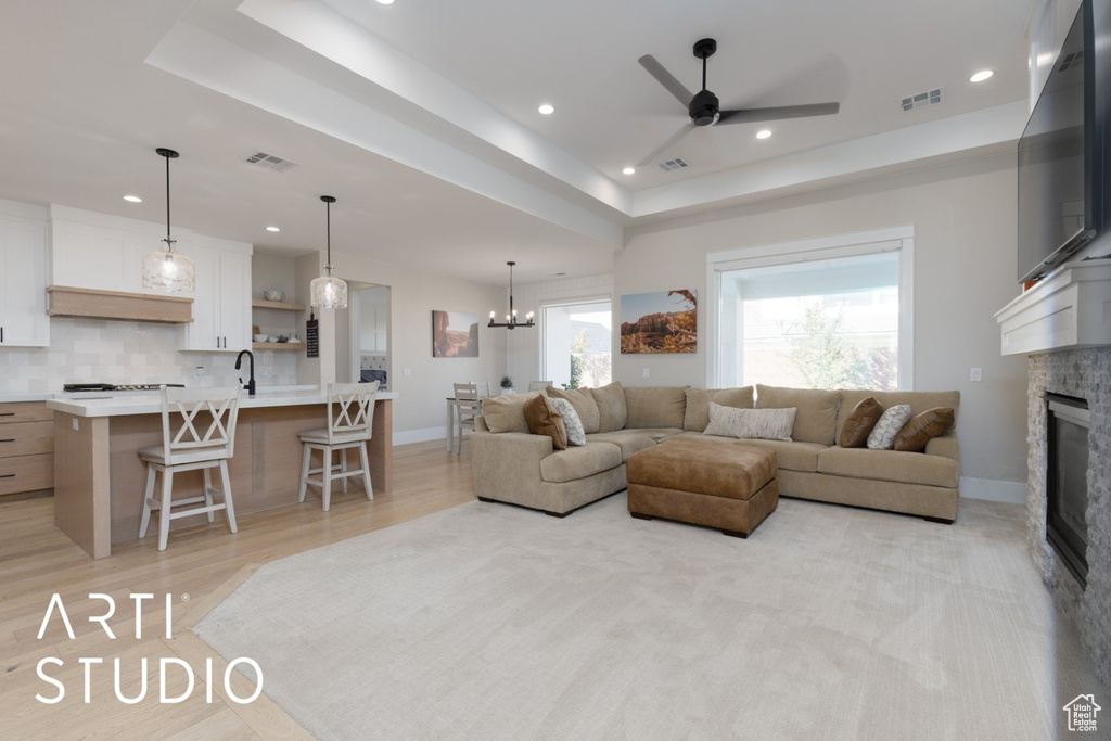 Living room with ceiling fan with notable chandelier, a stone fireplace, light wood-type flooring, and a raised ceiling