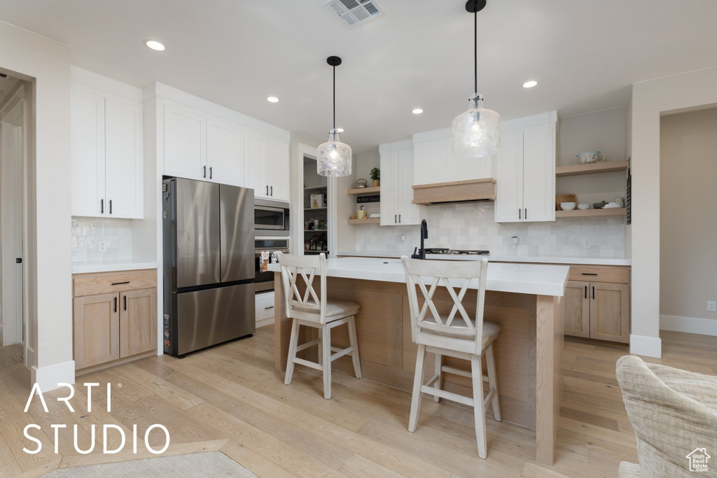 Kitchen featuring light wood-type flooring, stainless steel appliances, white cabinetry, a center island with sink, and backsplash