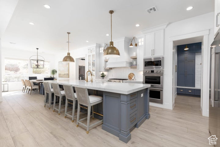 Kitchen with light wood-type flooring, stainless steel appliances, hanging light fixtures, a large island, and white cabinetry