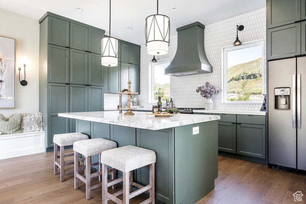 Kitchen with light stone counters, hanging light fixtures, stainless steel fridge, wall chimney exhaust hood, and dark hardwood / wood-style floors