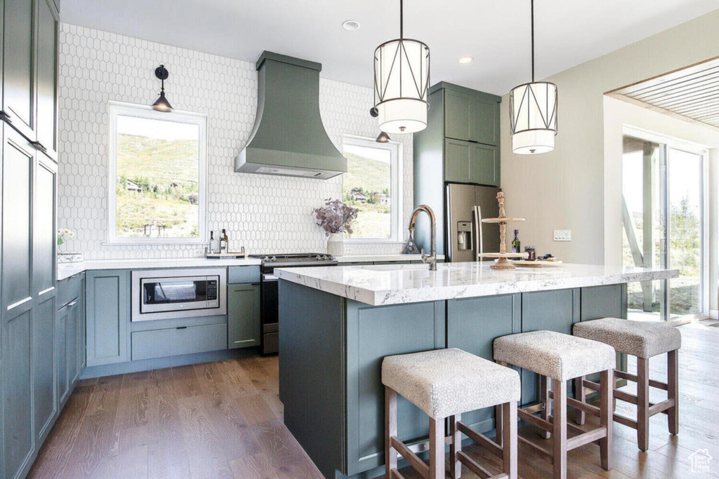 Kitchen with appliances with stainless steel finishes, a kitchen island with sink, wall chimney range hood, and a healthy amount of sunlight