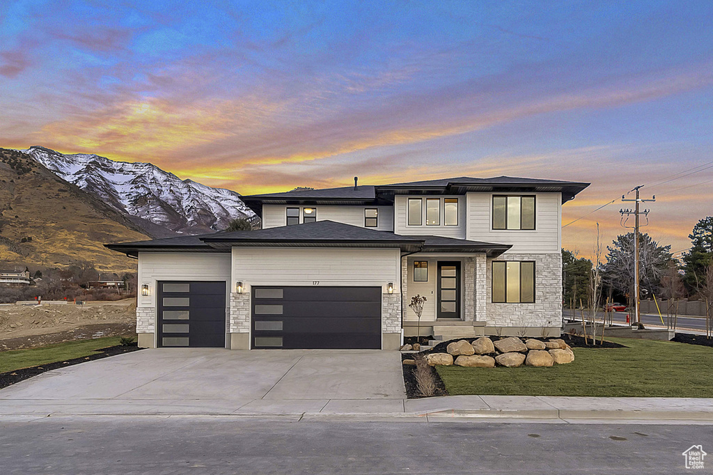 View of front of property with an attached garage, concrete driveway, stone siding, a lawn, and a mountain view