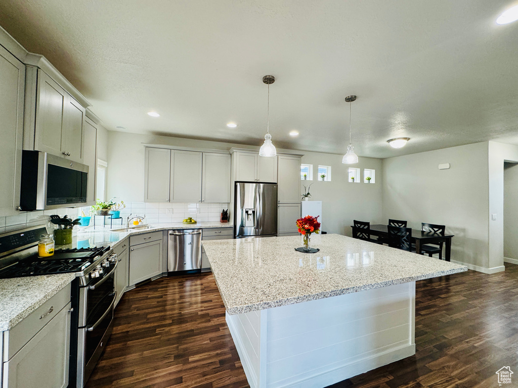 Kitchen with pendant lighting, dark wood-type flooring, appliances with stainless steel finishes, a center island, and light stone countertops