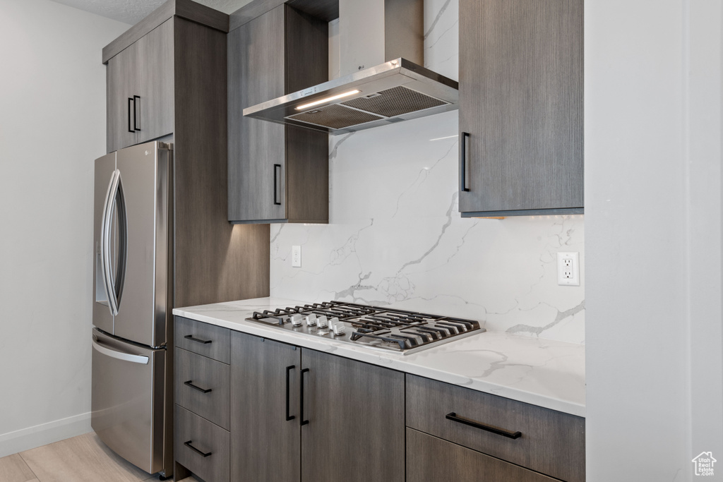 Kitchen featuring light wood-type flooring, light stone countertops, stainless steel appliances, tasteful backsplash, and wall chimney exhaust hood