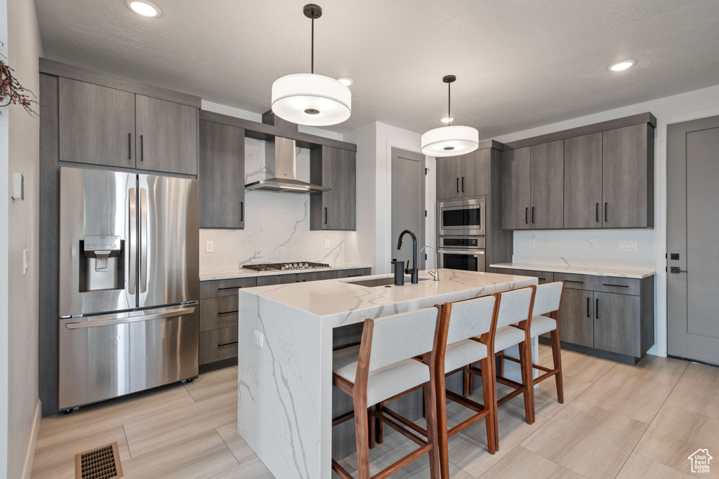 Kitchen featuring wall chimney exhaust hood, a center island with sink, decorative light fixtures, sink, and appliances with stainless steel finishes