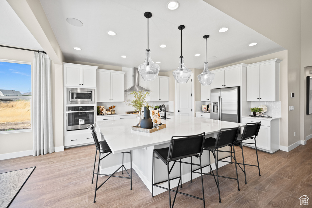 Kitchen featuring a large island with sink, stainless steel appliances, white cabinets, wall chimney exhaust hood, and light hardwood / wood-style floors