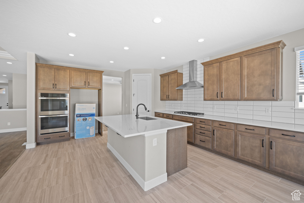 Kitchen featuring decorative backsplash, stainless steel appliances, wall chimney exhaust hood, sink, and a center island with sink