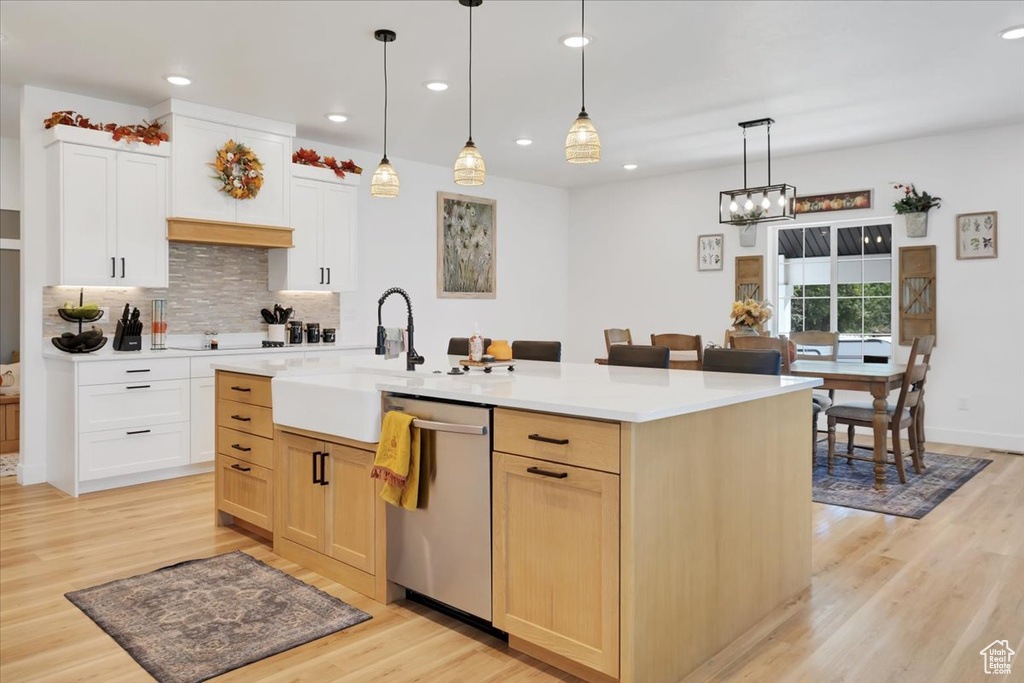 Kitchen with light wood-type flooring, a center island with sink, decorative light fixtures, stainless steel dishwasher, and white cabinetry