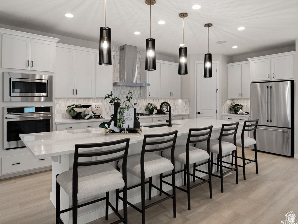 Kitchen featuring wall chimney exhaust hood, white cabinets, appliances with stainless steel finishes, and a kitchen island with sink