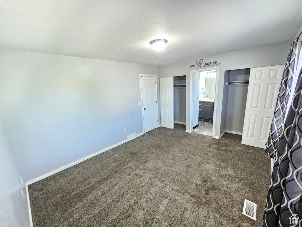 Unfurnished bedroom with two closets, dark carpet, and ensuite bath