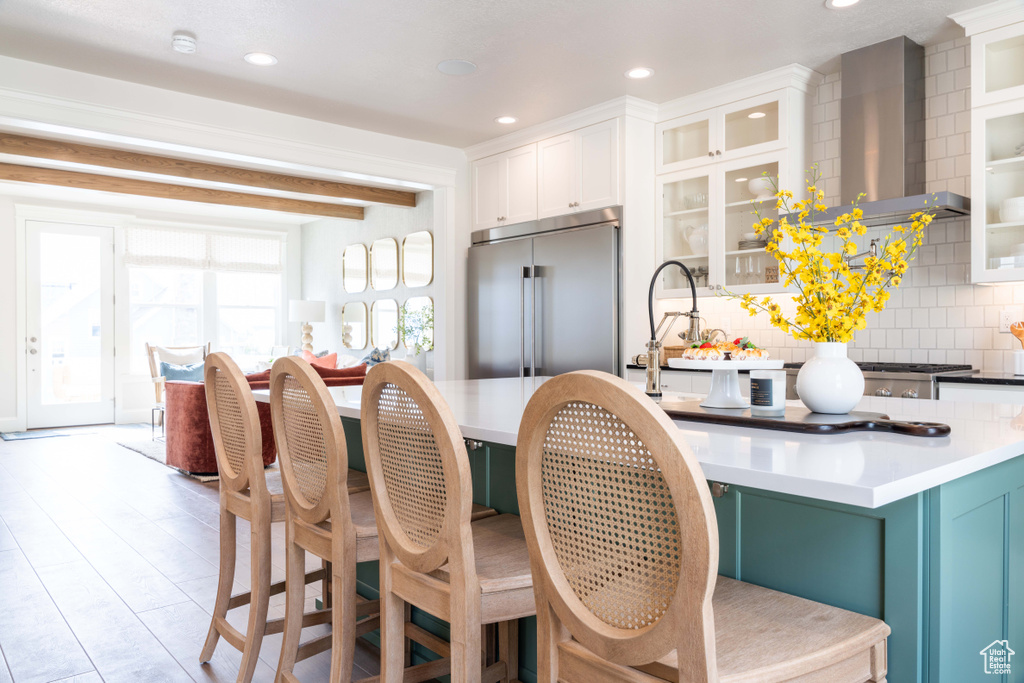 Kitchen with beamed ceiling, decorative backsplash, wall chimney exhaust hood, white cabinetry, and high end refrigerator