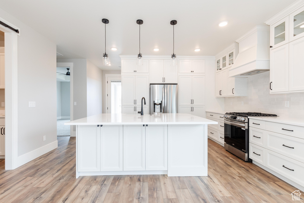 Kitchen featuring light hardwood / wood-style floors, pendant lighting, custom exhaust hood, an island with sink, and appliances with stainless steel finishes